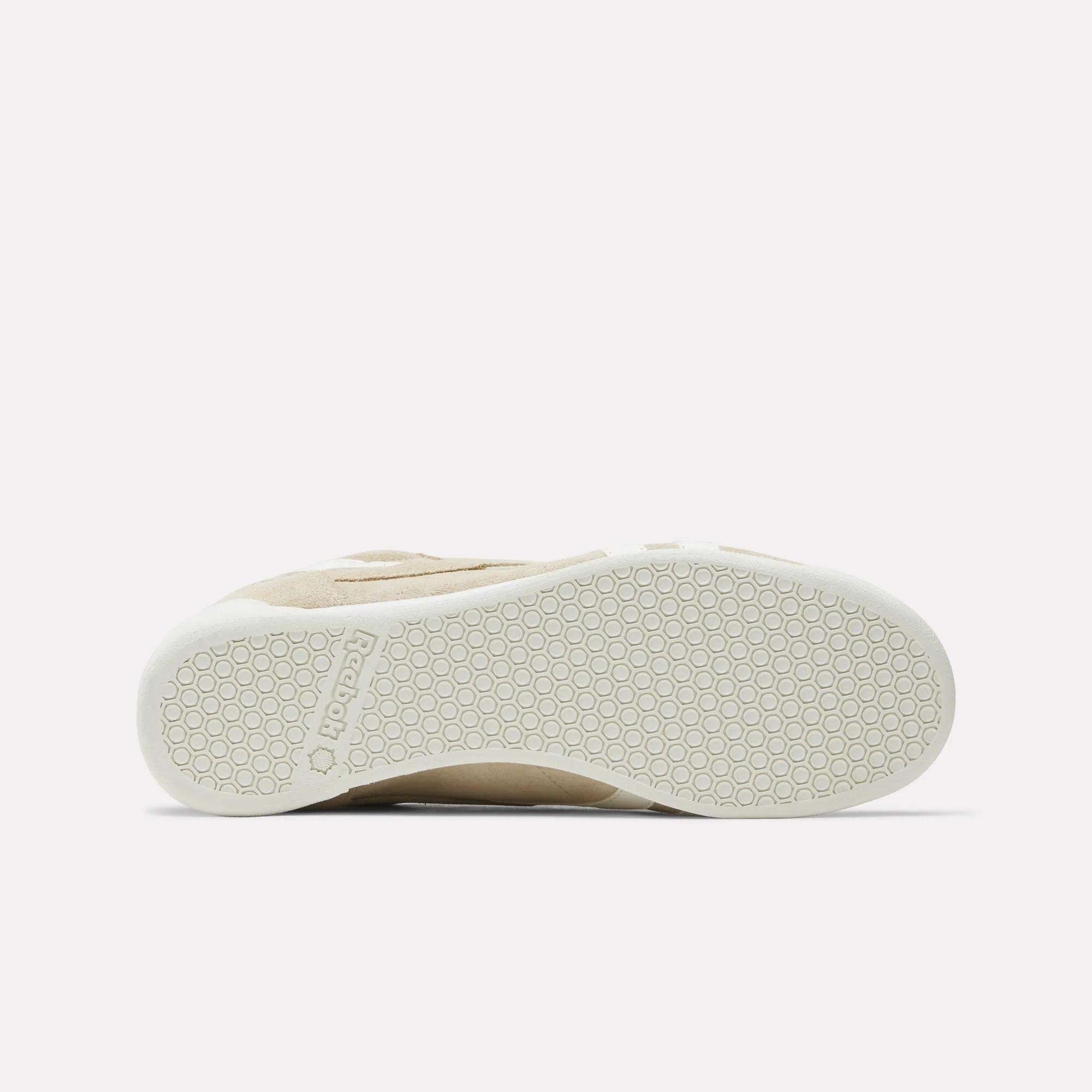 The image shows the bottom view of the Women's Freestyle Lo Shoes by Reebok, featuring a beige suede upper, a white rubber sole with a hexagonal pattern, and "Reebok" embossed near the heel.