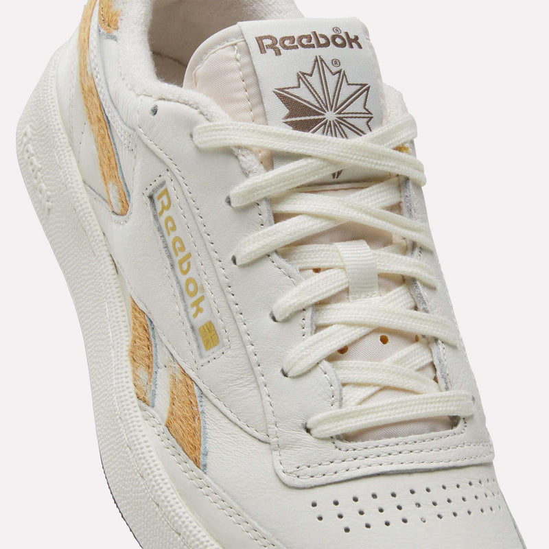 Close-up of the Reebok Club C Revenge Vintage Shoes, featuring a crisp white leather upper accented with tan and gold details. The shoe showcases the iconic Reebok logo alongside a maple leaf design on the tongue, while white laces threaded through the eyelets emphasize its classic tennis-inspired aesthetic.