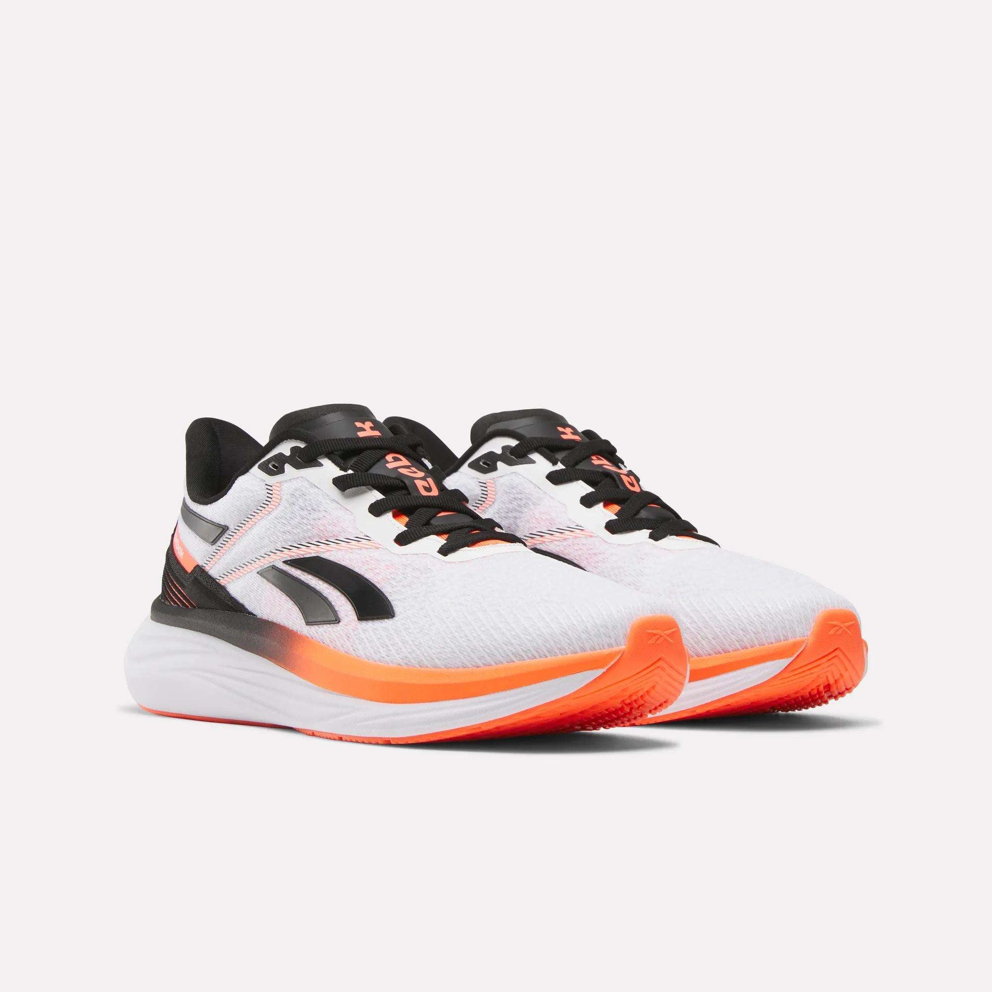 The Reebok Men's Viva Speed Shoes feature a white athletic design with black and orange accents, a thick cushioned sole, black laces, and a stylized logo on the side. Displayed against a plain white background.