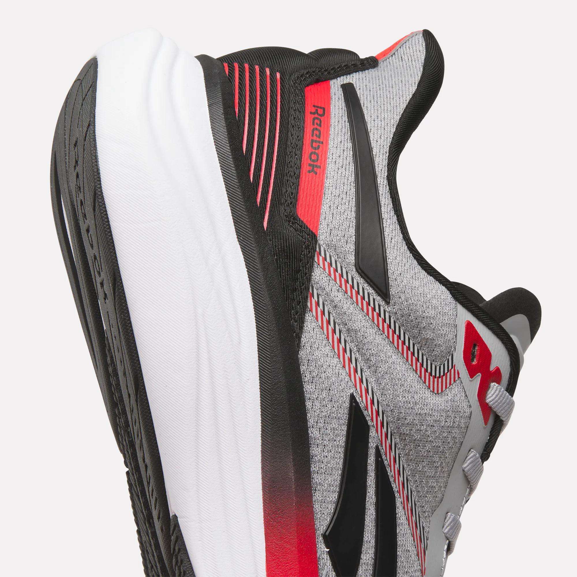 Close-up of Reebok Men's Viva Speed Shoes in grey, black, and red with a breathable mesh upper, textured fabric, cushioned white and black sole, and Reebok logos on the side and heel.