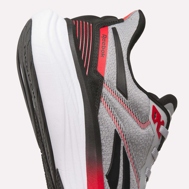 Close-up of Reebok Men's Viva Speed Shoes in grey, black, and red with a breathable mesh upper, textured fabric, cushioned white and black sole, and Reebok logos on the side and heel.