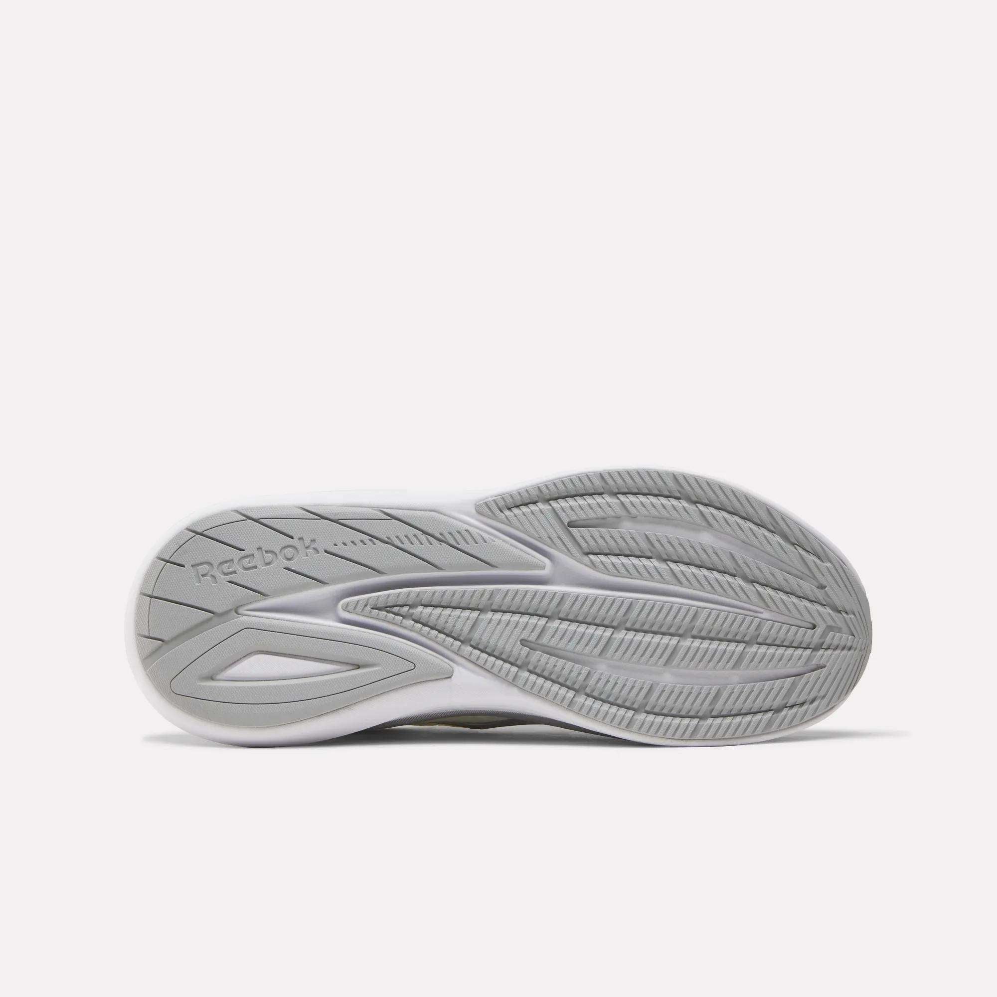 The image displays the sole of Reebok Men's Viva Speed Shoes in white and gray, featuring textured tread patterns, grooves for traction, and the Reebok logo on the left side, emphasizing the shoes’ enhanced cushioning.