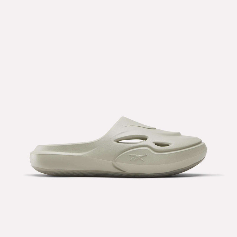 The Reebok DMX Recovery Mules are light beige slide sandals with a thick molded EVA sole, upper cutout details, and an embossed Reebok logo, shown in side profile against a plain white background.