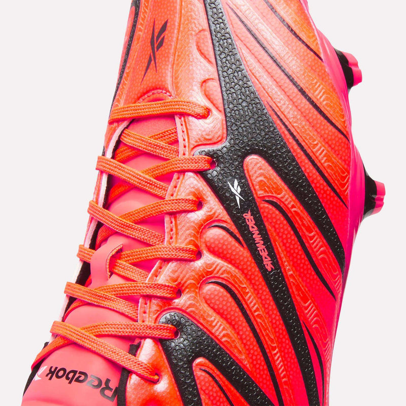 Close-up of the Reebok Sidewinder 26 League Firm Ground Cleats in bright orange and black, featuring textured patterns, orange laces, and the Reebok logo on both the side and tongue. Engineered for optimal ball control, these soccer cleats are equipped with firm ground studs to provide superior traction on natural grass surfaces.