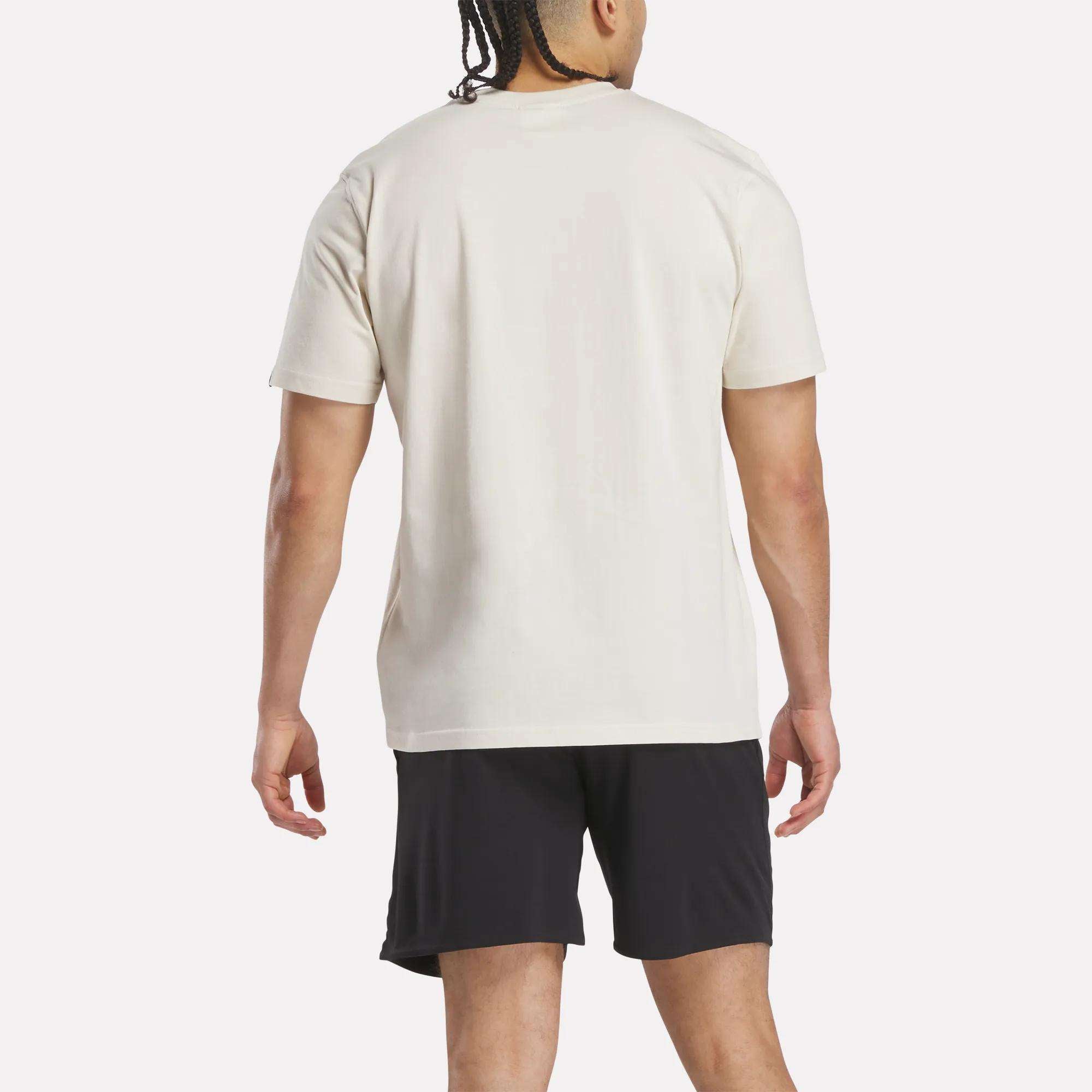 A man with braided hair faces away from the camera, wearing a Reebok Men's Club Graphic T-Shirt in plain white and black shorts, set against a light background.