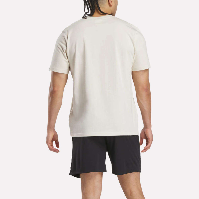 A man with braided hair faces away from the camera, wearing a Reebok Men's Club Graphic T-Shirt in plain white and black shorts, set against a light background.