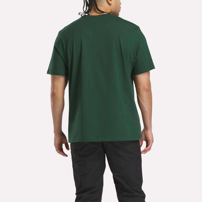 A person with braided hair is shown from the back, wearing a Reebok Men's International Graphic T-Shirt in dark green and black pants, standing against a plain light background.
