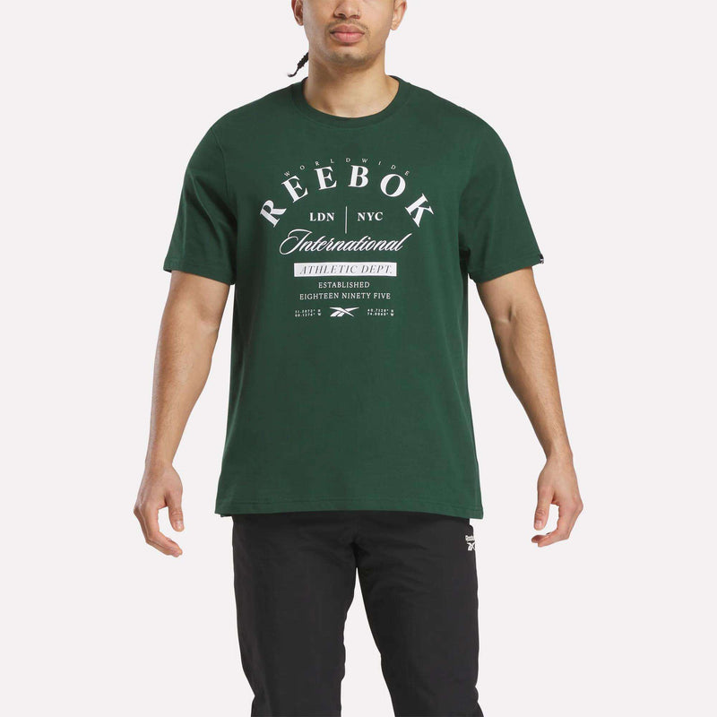 A man wears the Reebok Men's International Graphic T-Shirt in dark green with white graphics, paired with black pants and set against a plain white background, for a comfortable, stylish everyday look.