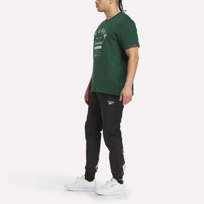 A person stands sideways against a white background, wearing a green Men's International Graphic T-Shirt by Reebok, black jogger pants with white Reebok logos, and white sneakers.