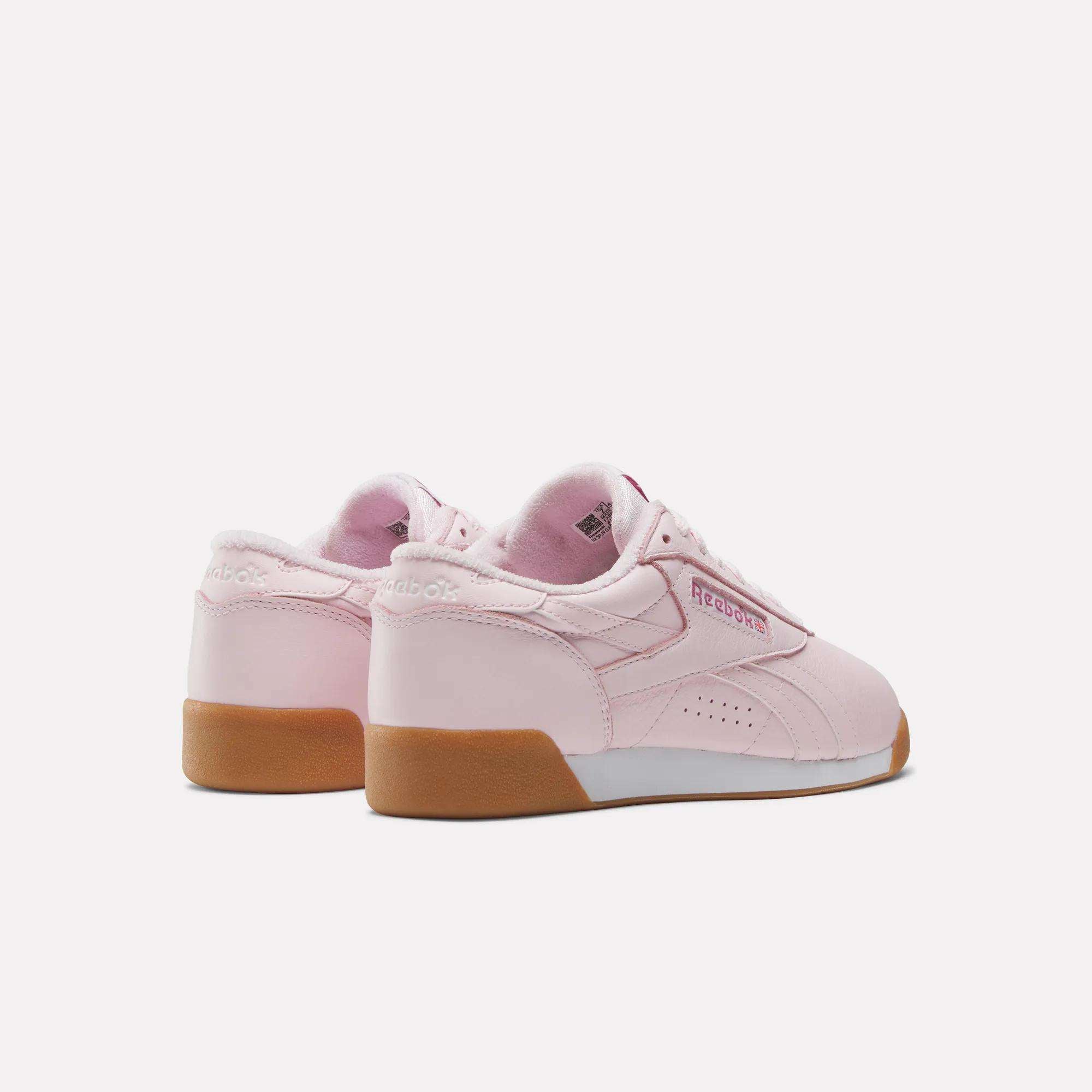 A pair of light pink Reebok Women's Freestyle Lo Shoes with gum-colored soles are shown from the back and side on a white background. The Reebok logo is visible on the heel and side, making them ideal for everyday wear.
