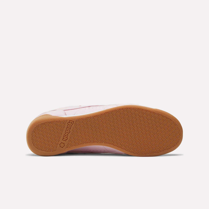 The image displays the bottom view of Reebok Women's Freestyle Lo Shoes, featuring a light brown rubber sole with a hexagonal pattern and a stylish pink and white upper, perfect for everyday wear.