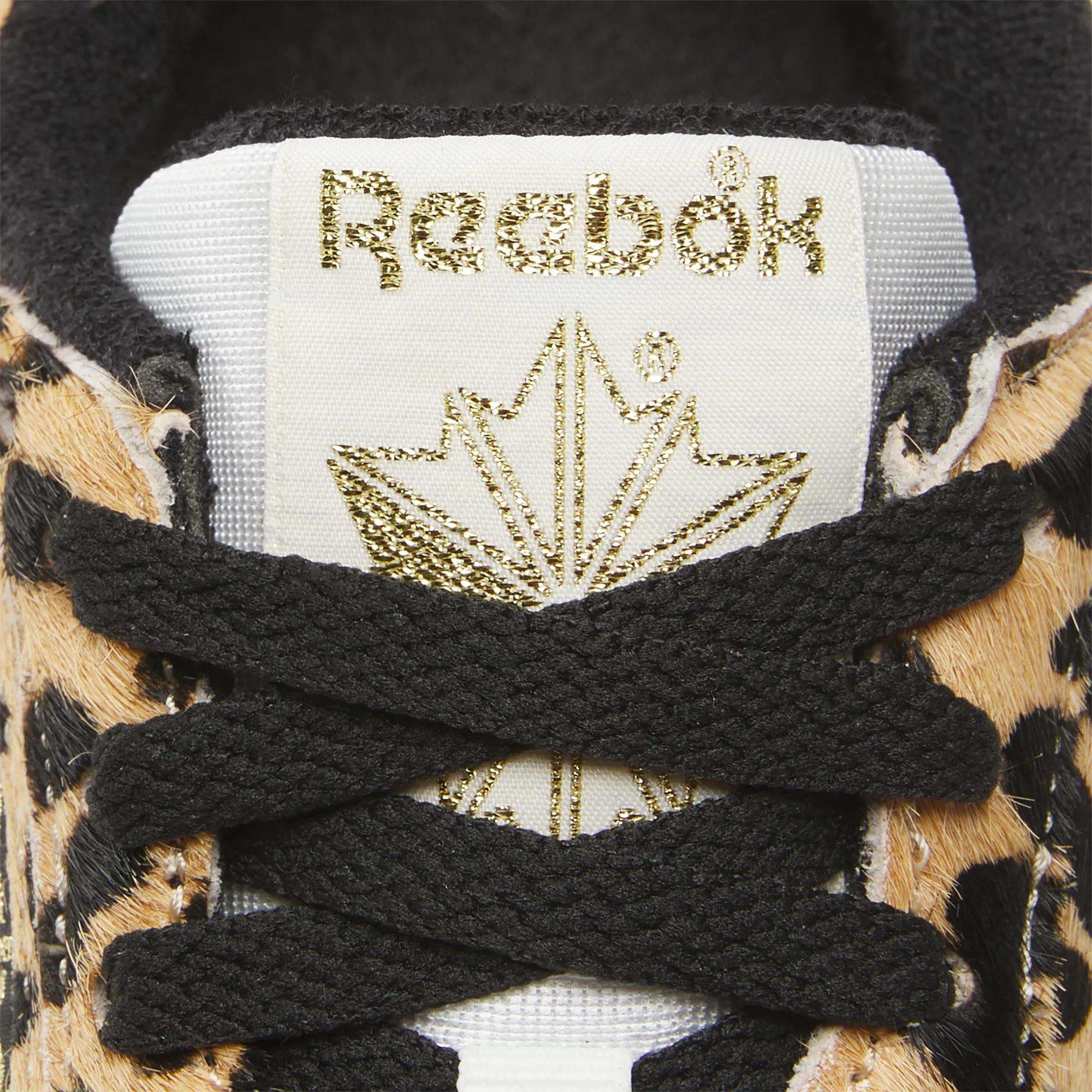 Close-up of the Reebok Women's Freestyle Lo Shoes shows a gold logo and maple leaf on the tongue, black laces, and tan/black animal print pony hair upper—an eye-catching nod to classic '80s fitness trainers.