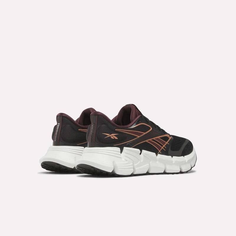 A pair of Women's FloatZig 2 Running Shoes by Reebok, black with orange accents and thick white soles featuring Superfloat+ cushioning, are shown on a white background facing backward and slightly to the side.
