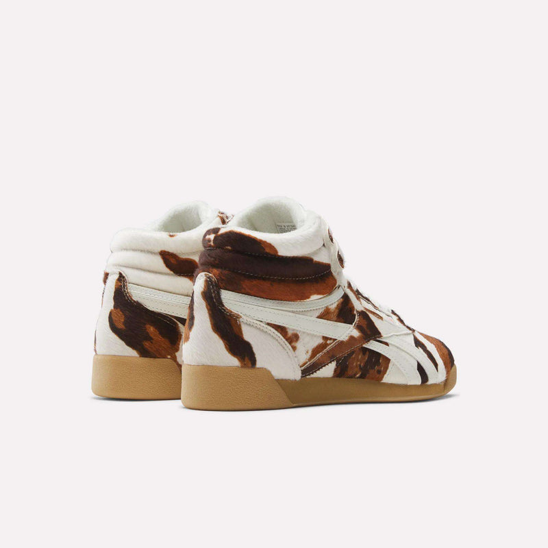 The Reebok Women's Freestyle Hi Shoes are '80s-inspired high tops with a brown and white cowhide pattern, beige gum soles, and white accents. Shown from the back and side against a plain white background.