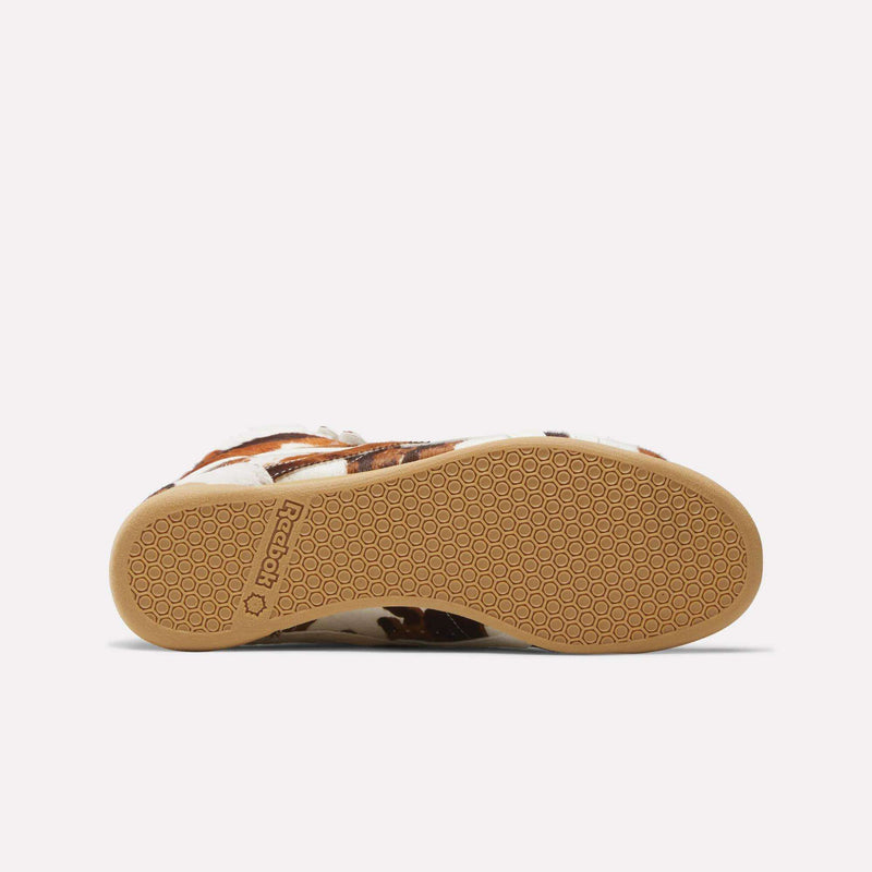 A close-up of the outsole of the Reebok Women's Freestyle Hi Shoes, a classic '80s fitness style, reveals a honeycomb-patterned light brown rubber sole with the Reebok logo near the heel and part of the brown and white upper visible.
