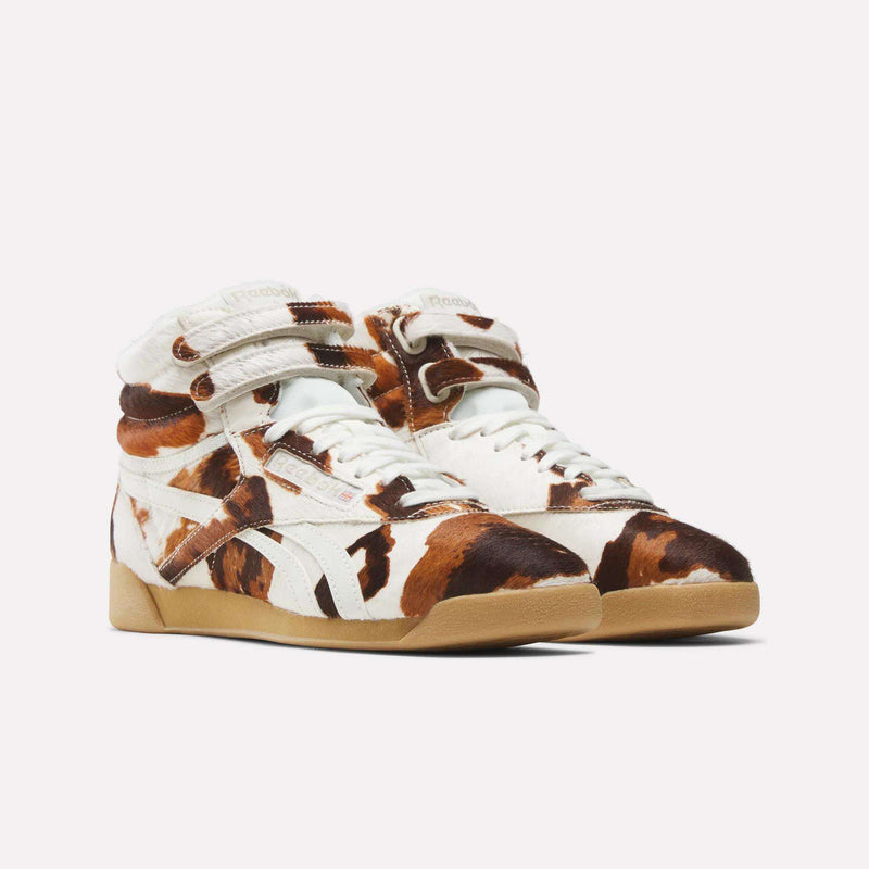 The Women's Freestyle Hi Shoes by Reebok feature a brown and white cow print, white laces, and light brown rubber soles, evoking '80s fitness style. Shown against a plain white background.