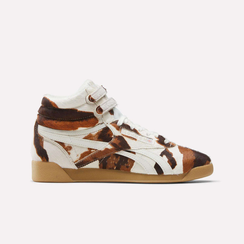 The Reebok Women's Freestyle Hi Shoes are high-top sneakers inspired by '80s classics, featuring a brown, white, and black cowhide pattern, two white Velcro ankle straps, white laces, and a light brown rubber sole.