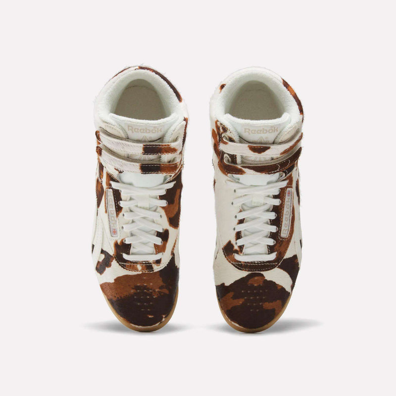 The Women's Freestyle Hi Shoes by Reebok feature a bold brown and white cowhide pattern, white laces, and white soles, viewed from above—embodying the iconic spirit of this '80s fitness classic.