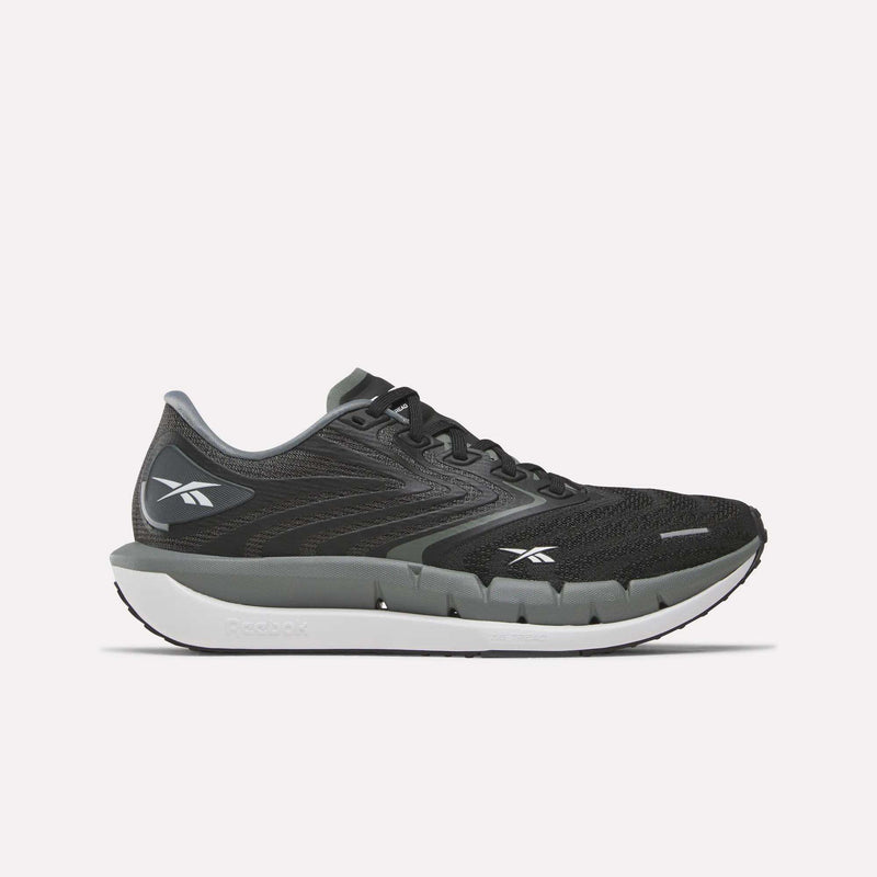 A pair of Reebok Women’s FloatZig Tread Running Shoes in Black/Washed Black/Chalk, featuring a white sole, the Reebok logo on the side and heel, and a breathable mesh upper, is shown from the left against a plain white background.