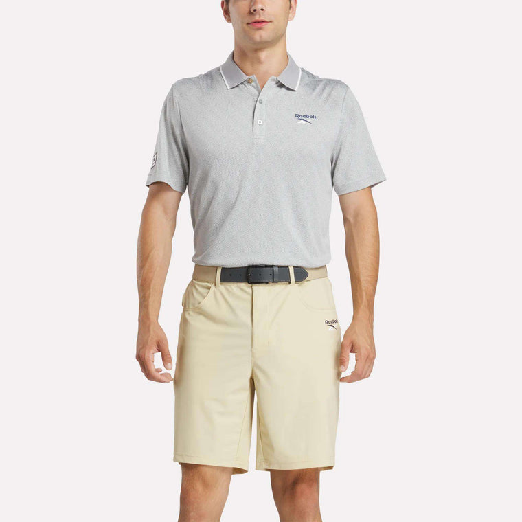 A man wears the Reebok Men's BDC Signature Polo Shirt in light gray, made with UPF-rated RBK-DRY fabric, tucked into beige shorts with a black belt, standing against a plain white background.