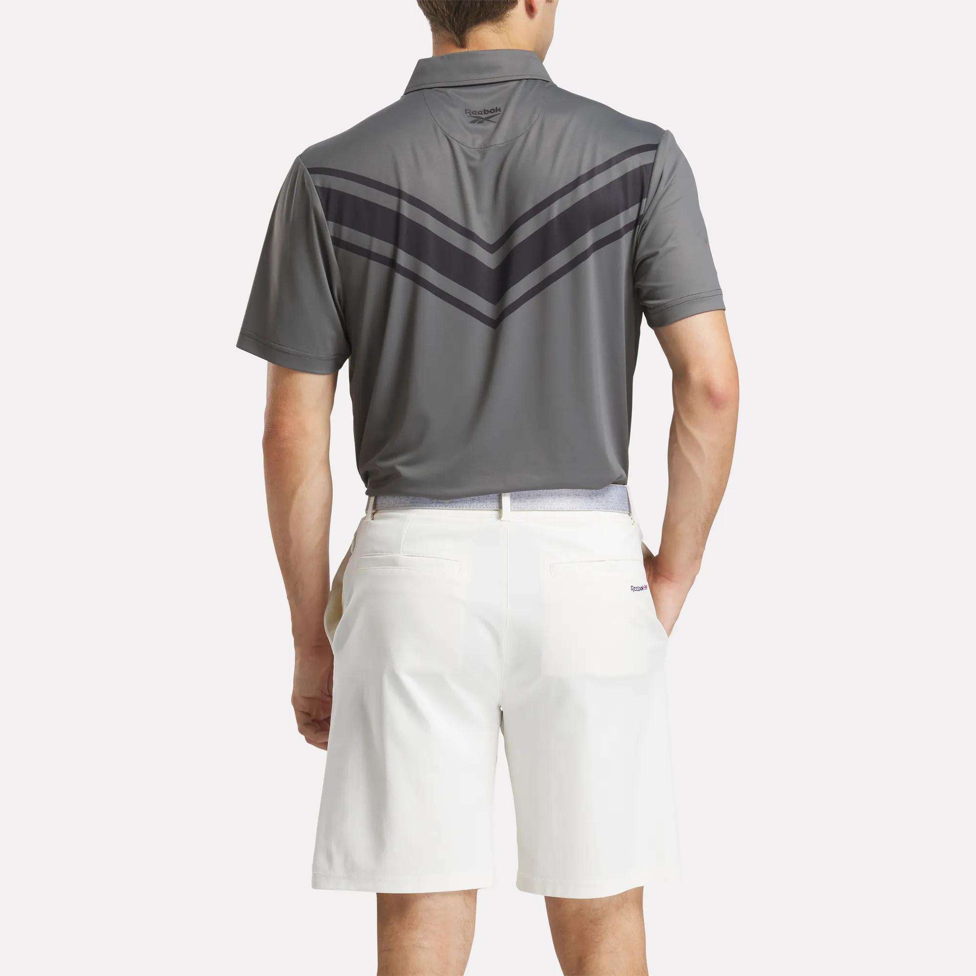 A man stands with his back to the camera, wearing a gray Reebok Men's BDC Colorblocked Polo Shirt featuring RBK-DRY tech and dark chevron stripes, paired with white shorts, against a plain background.