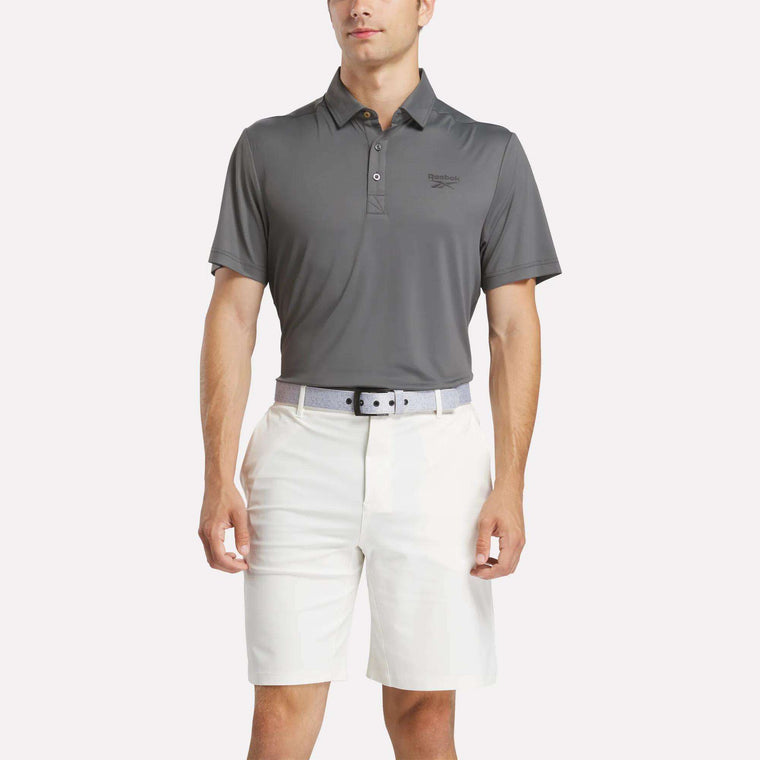 A man in a Men's BDC Colorblocked Polo Shirt by Reebok, tucked into white shorts with a light gray belt, stands against a plain backdrop, highlighting classic Reebok men's style.