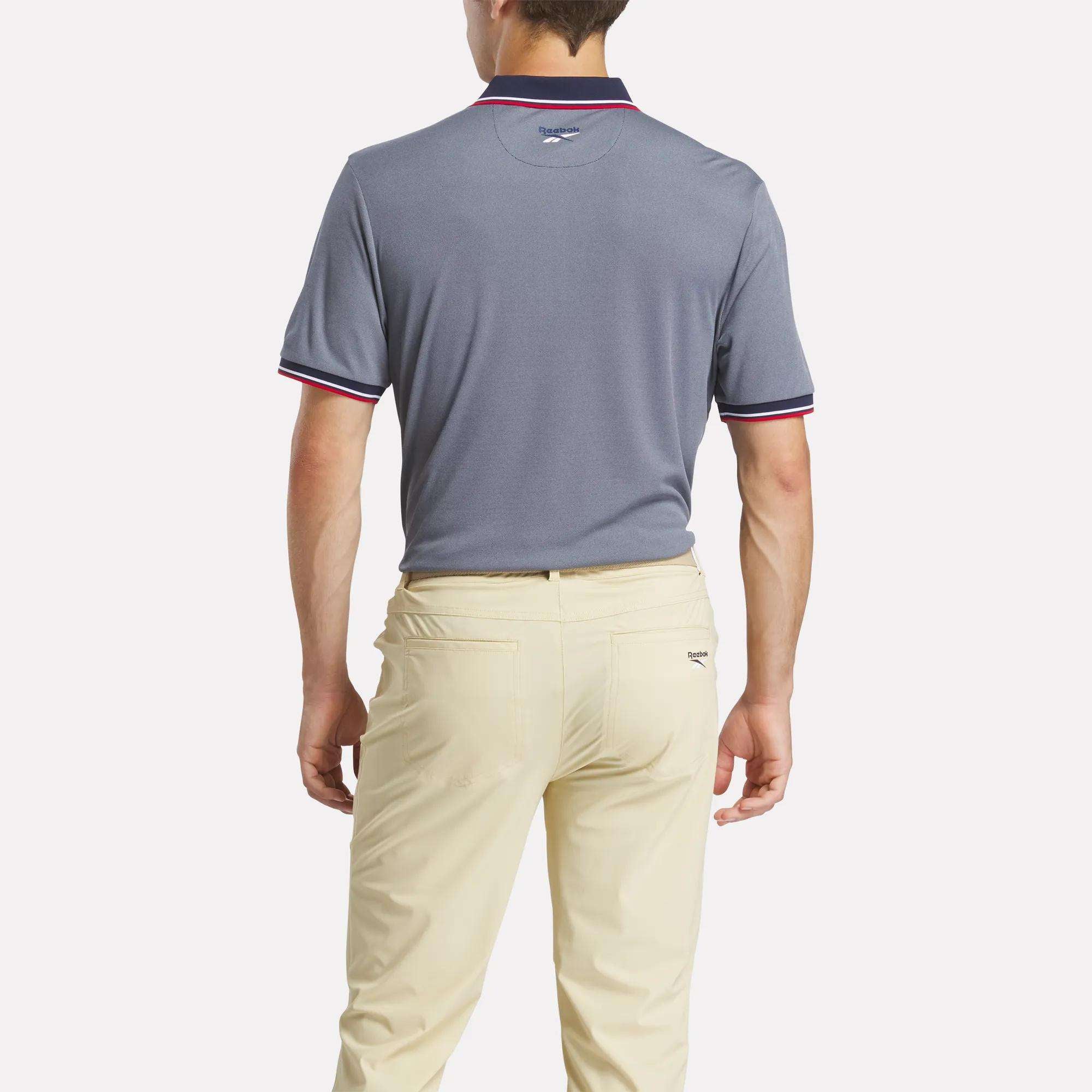 A man, shown from the back, wears a gray Reebok Men's Country Club Oxford Pique Polo Shirt with red and white trim and light beige pants, standing against a plain white background.