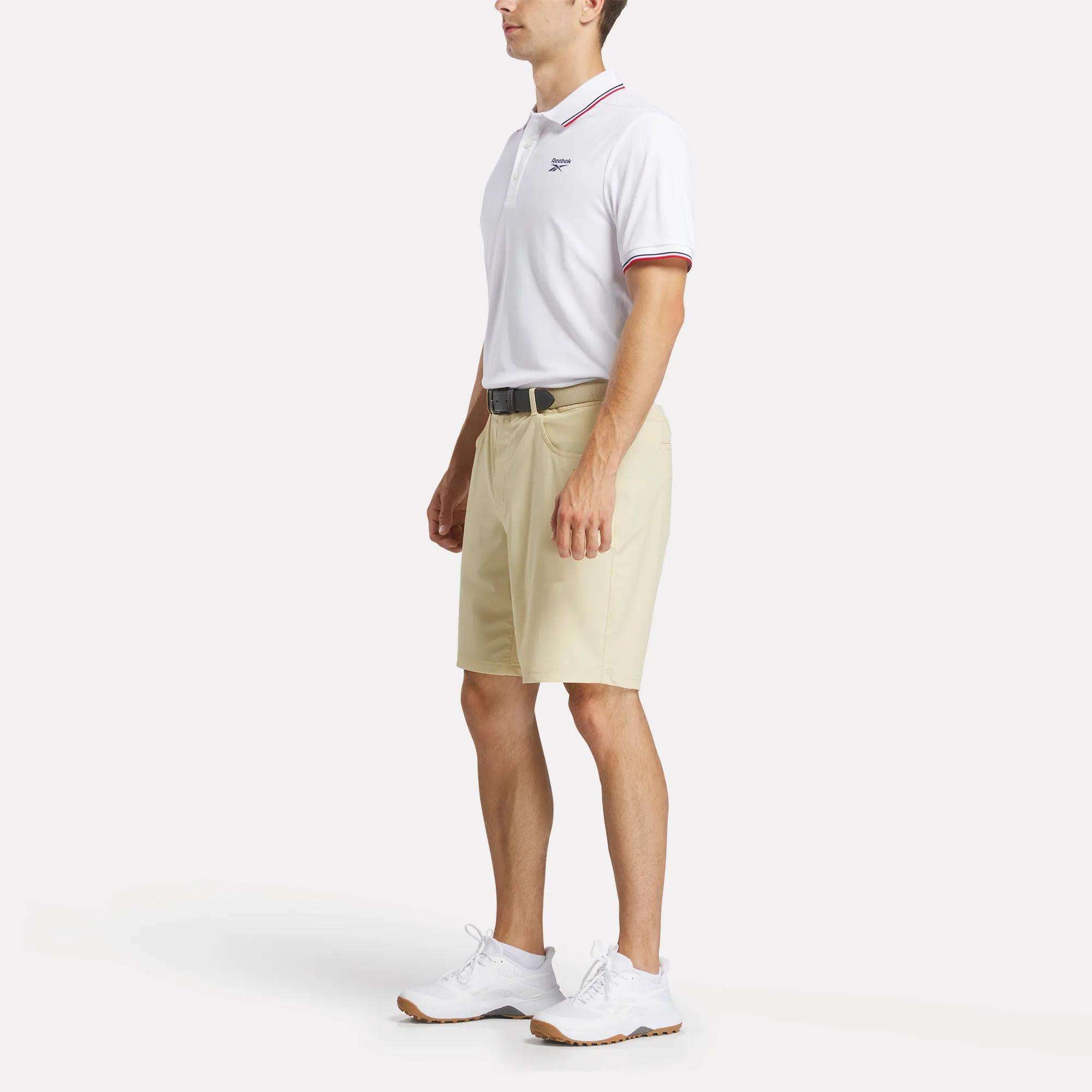 A man stands against a plain light background wearing a Reebok Men's Country Club Oxford Pique Polo Shirt, featuring red trim, paired with beige knee-length shorts, a black belt, and white sneakers.