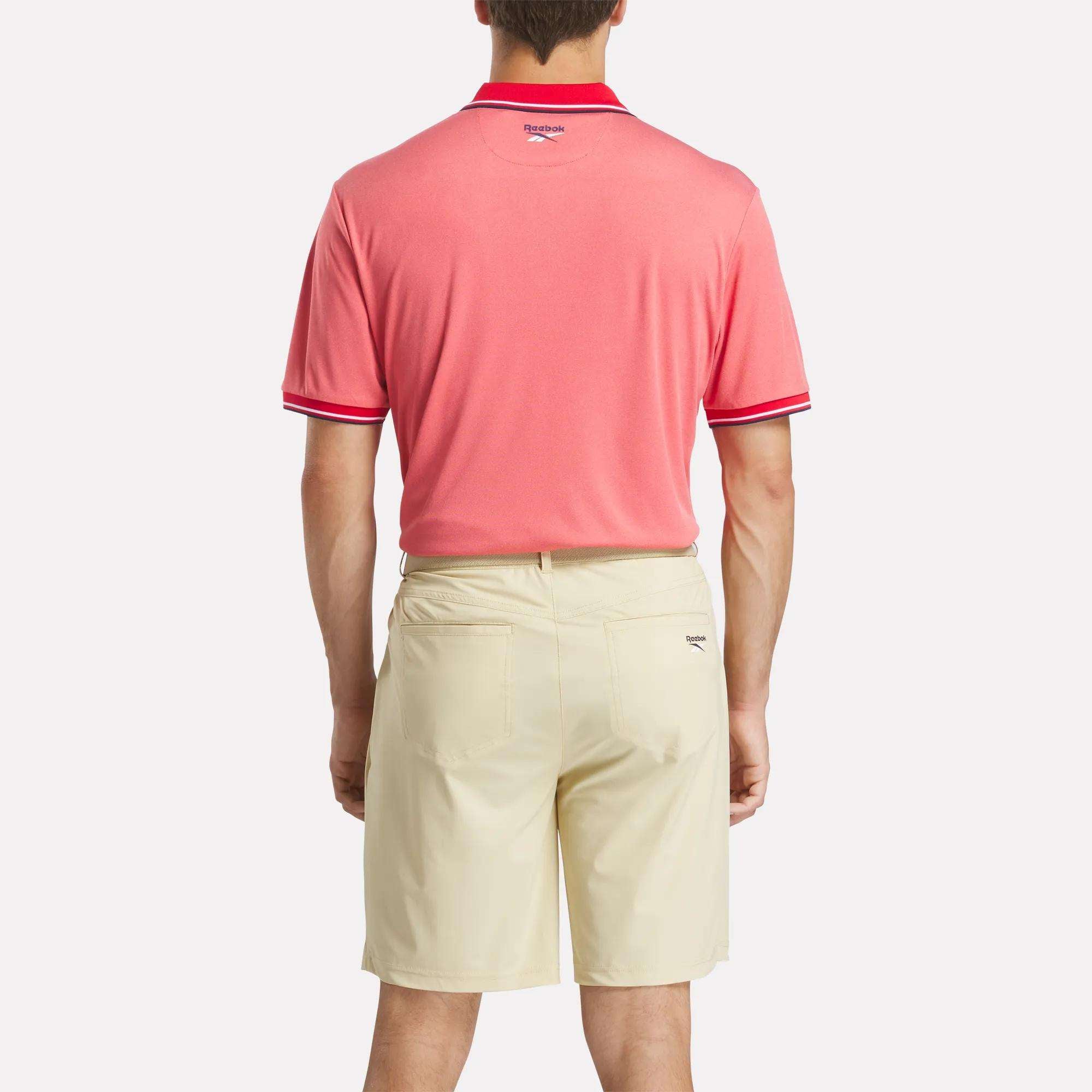 A man stands with his back to the camera wearing a pink Reebok Men's Country Club Oxford Pique Polo Shirt with red trim and light beige shorts, set against a plain white background.