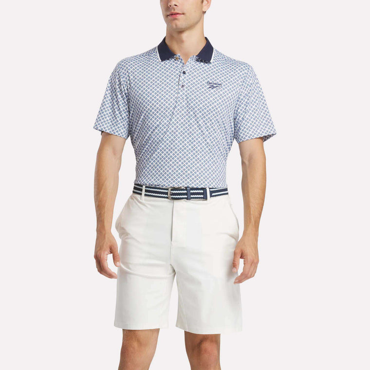 A man wears a Reebok Men's Sublimation Print Shirt with blue and white patterns, paired with white shorts and a matching belt, standing against a plain white background.