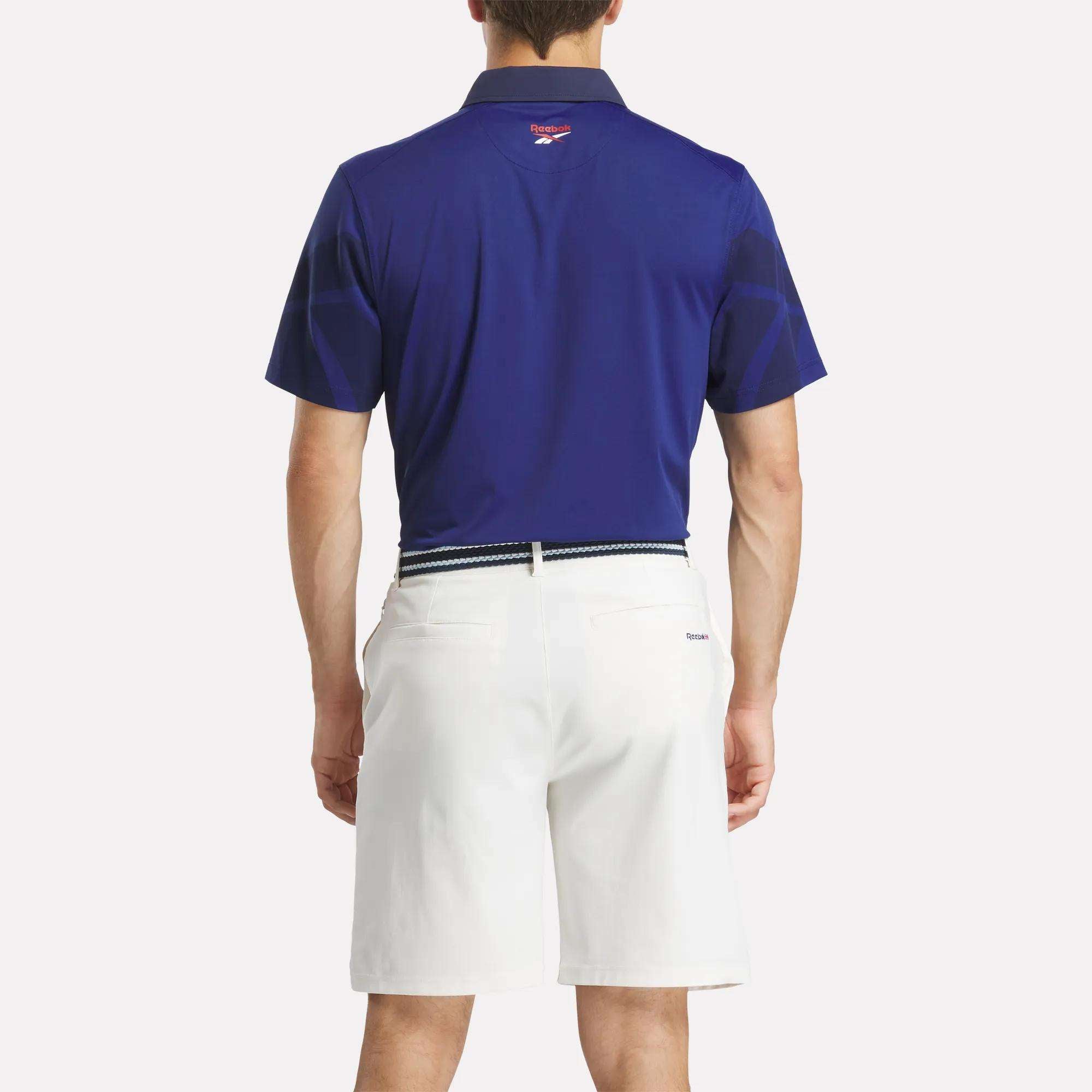 A man faces away, wearing the Reebok Men's Signature Vector Colorblocked Polo Shirt in dark blue, tucked into white shorts with a belt. The background is plain and light-colored.