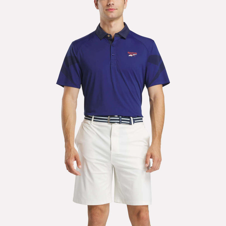 A man faces forward wearing the Reebok Men's Signature Vector Colorblocked Polo Shirt with UPF 30+, a small chest logo, white knee-length shorts, and a blue-and-white belt. The background is plain and light-colored.