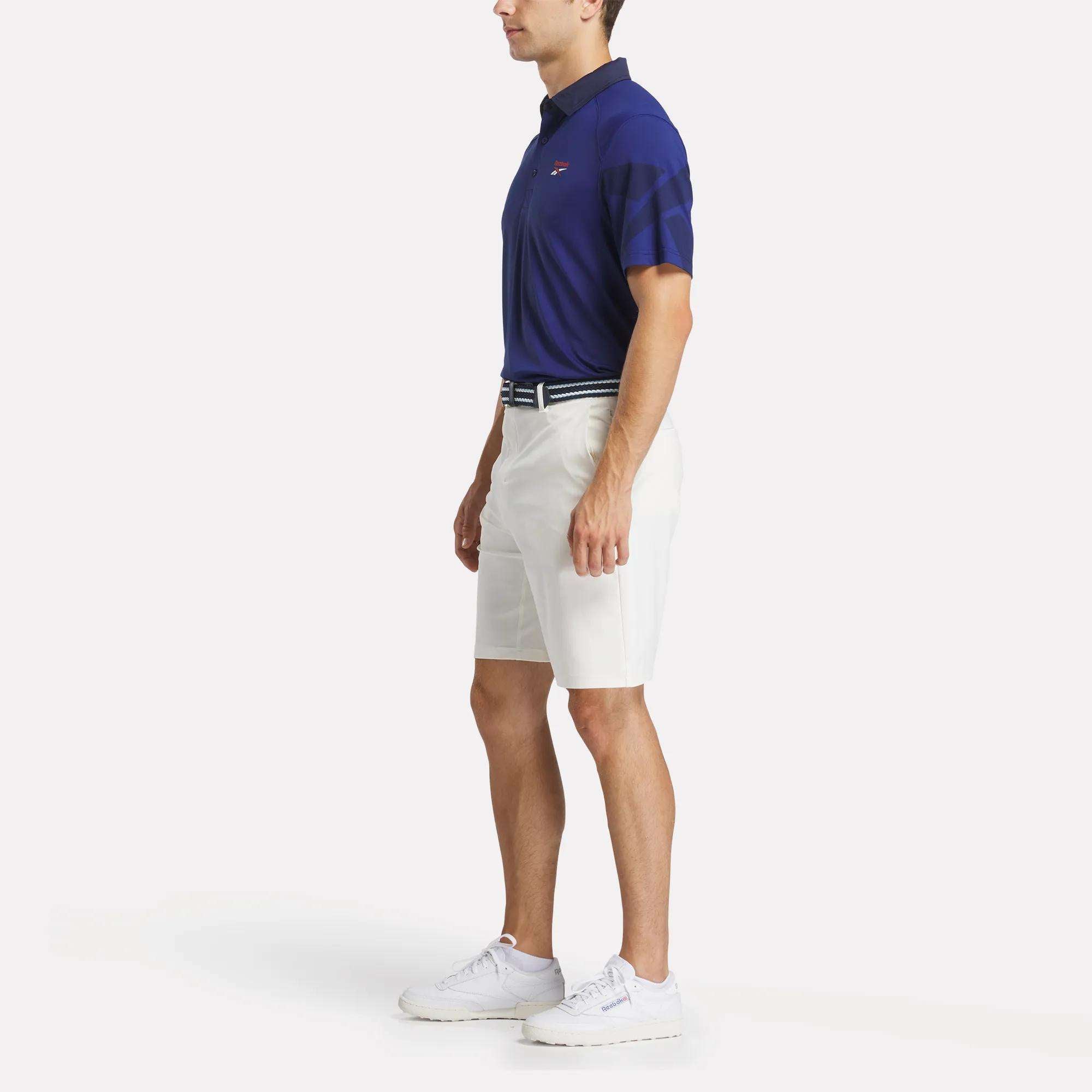 A man in profile wears the Reebok Men's Signature Vector Colorblocked Polo Shirt, white knee-length shorts, a striped belt, and white sneakers against a plain white background.