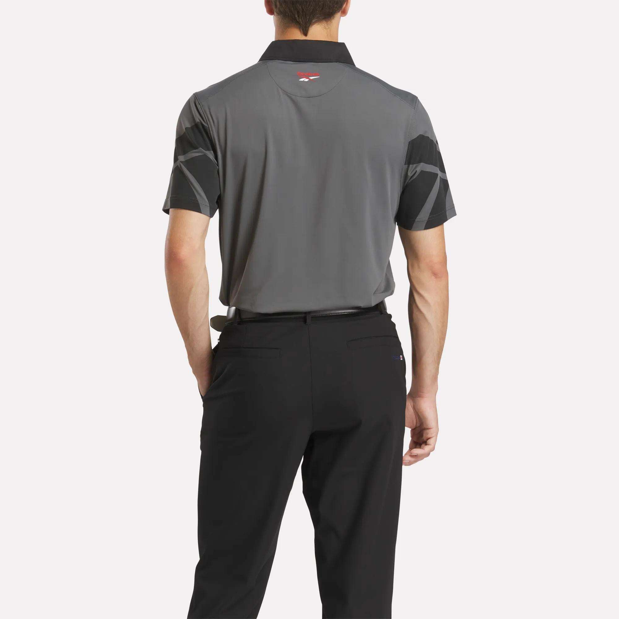 A man with his back to the camera wears a Reebok Men's Signature Vector Colorblocked Polo Shirt in gray and black, paired with black pants, set against a plain white background.