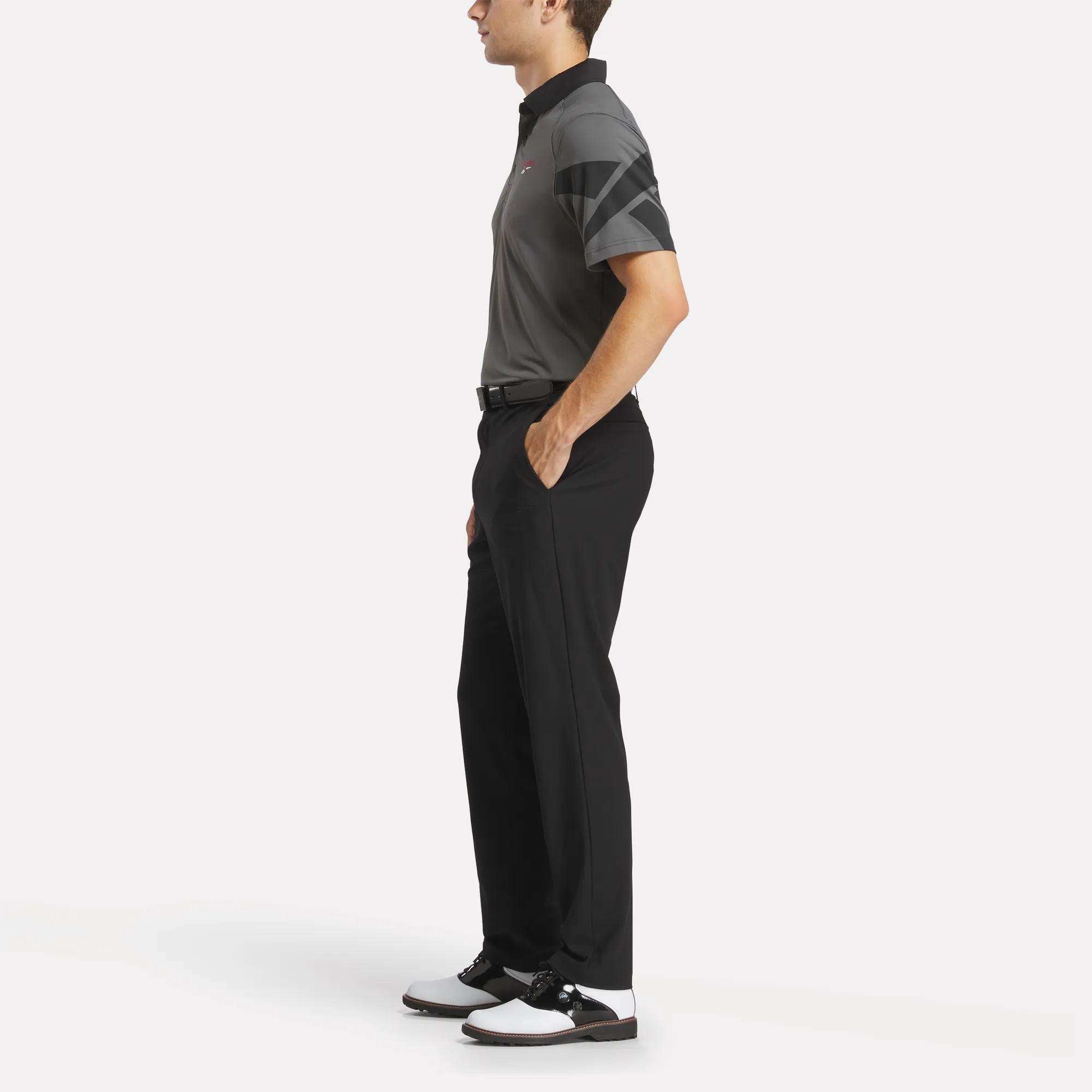 A man stands in profile with hands in his pockets, wearing a black and gray Reebok Men's Signature Vector Colorblocked Polo Shirt, black pants, and black and white golf shoes against a plain white background.