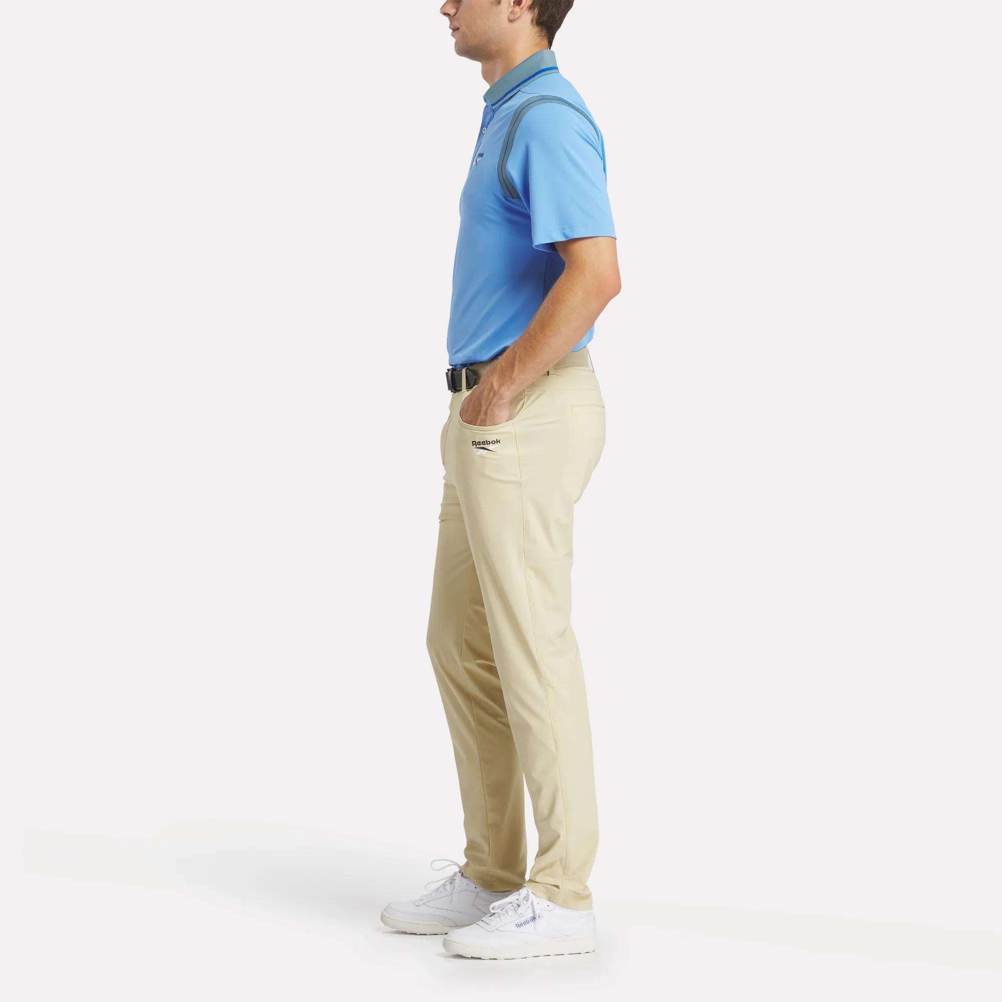 A man in profile wears a Reebok Men's Varsity Polo Shirt in light blue, beige pants, a black belt, and white sneakers, with hands in pockets against a plain white background.