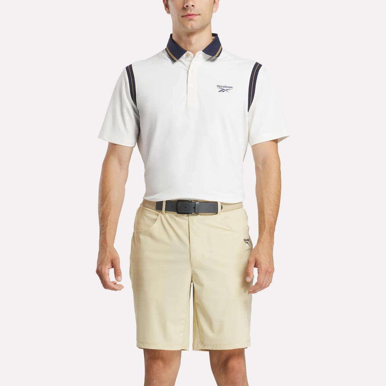 A man stands facing forward with hands relaxed, wearing a white Reebok Men's Varsity Polo Shirt with dark accents and beige shorts against a plain light background.