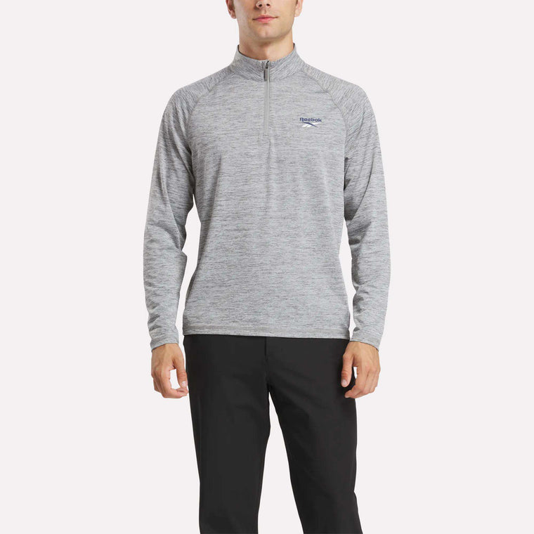 A man wearing a light gray Reebok Men's Cationic Wicking Quarter-Zip Sweatshirt with RBK-DRY tech and black pants stands against a plain white background.