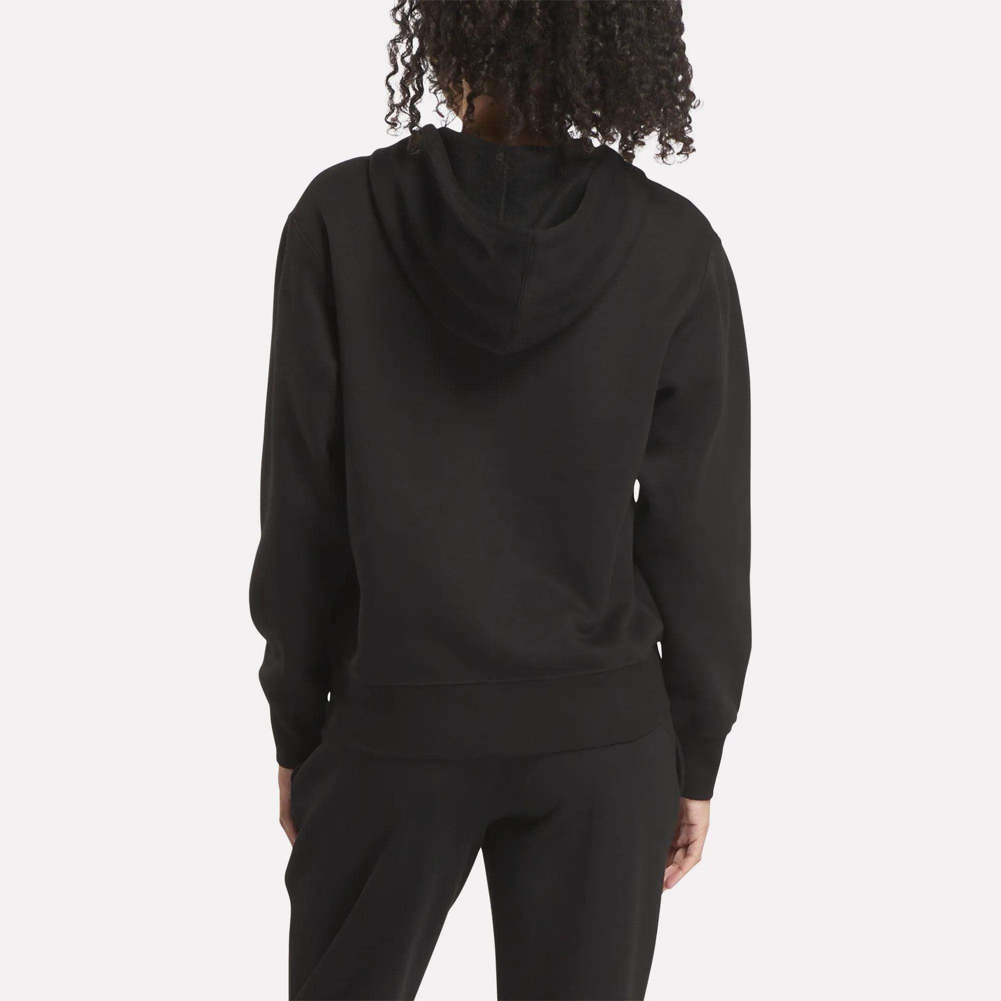 A person with curly hair is shown from the back, wearing a black Reebok Women's Small Vector Full Zip Sweatshirt and black pants, standing against a plain light background.