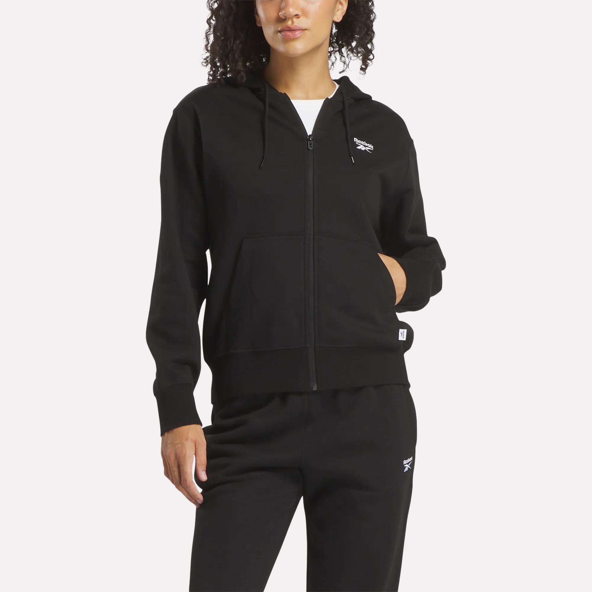 A person wearing a Reebok Women's Small Vector Full Zip Sweatshirt and black pants with small white logos, posing with one hand in the sweatshirt pocket against a plain light background.