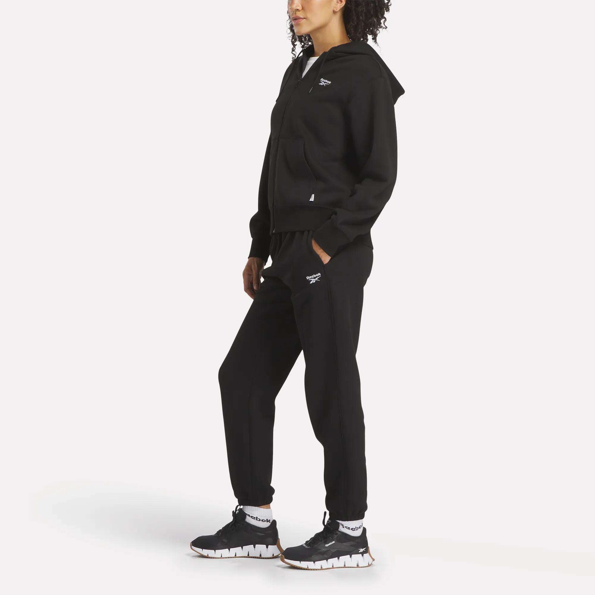 A woman poses in a relaxed stance wearing the Reebok Women's Small Vector Full Zip Sweatshirt in black, black sweatpants, black athletic shoes, and white socks against a plain white background.