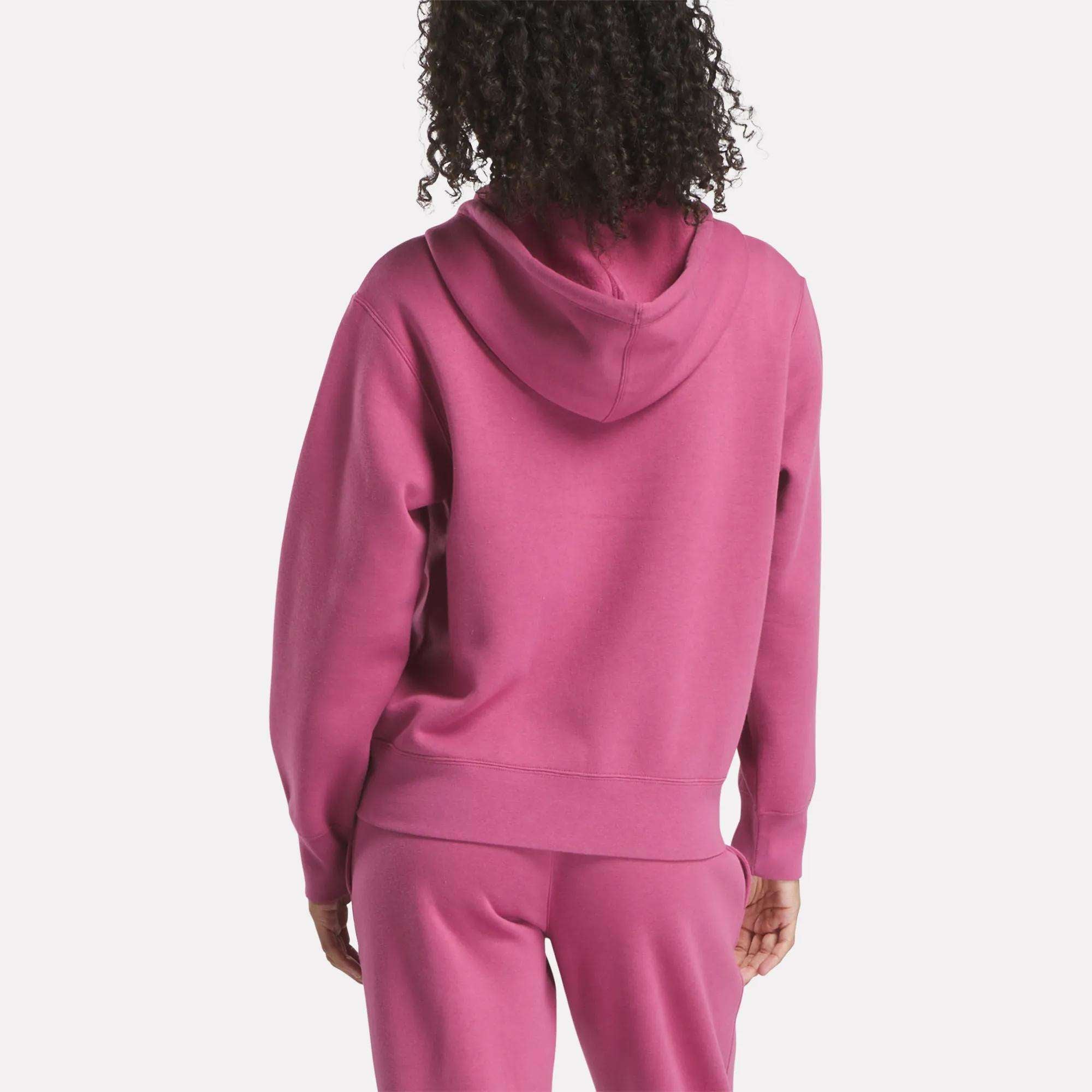 A woman with curly hair is seen from the back wearing a pink Reebok Women's Small Vector Full Zip Sweatshirt, paired with matching sweatpants against a plain white background.