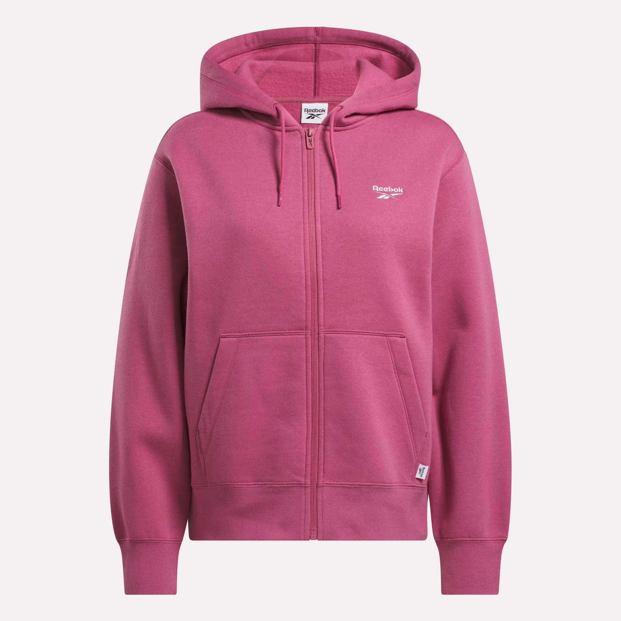 A pink Reebok Women's Small Vector Full Zip Sweatshirt features a drawstring hood, front pockets, and a small white Reebok logo on the left chest. It's shown against a plain white background.
