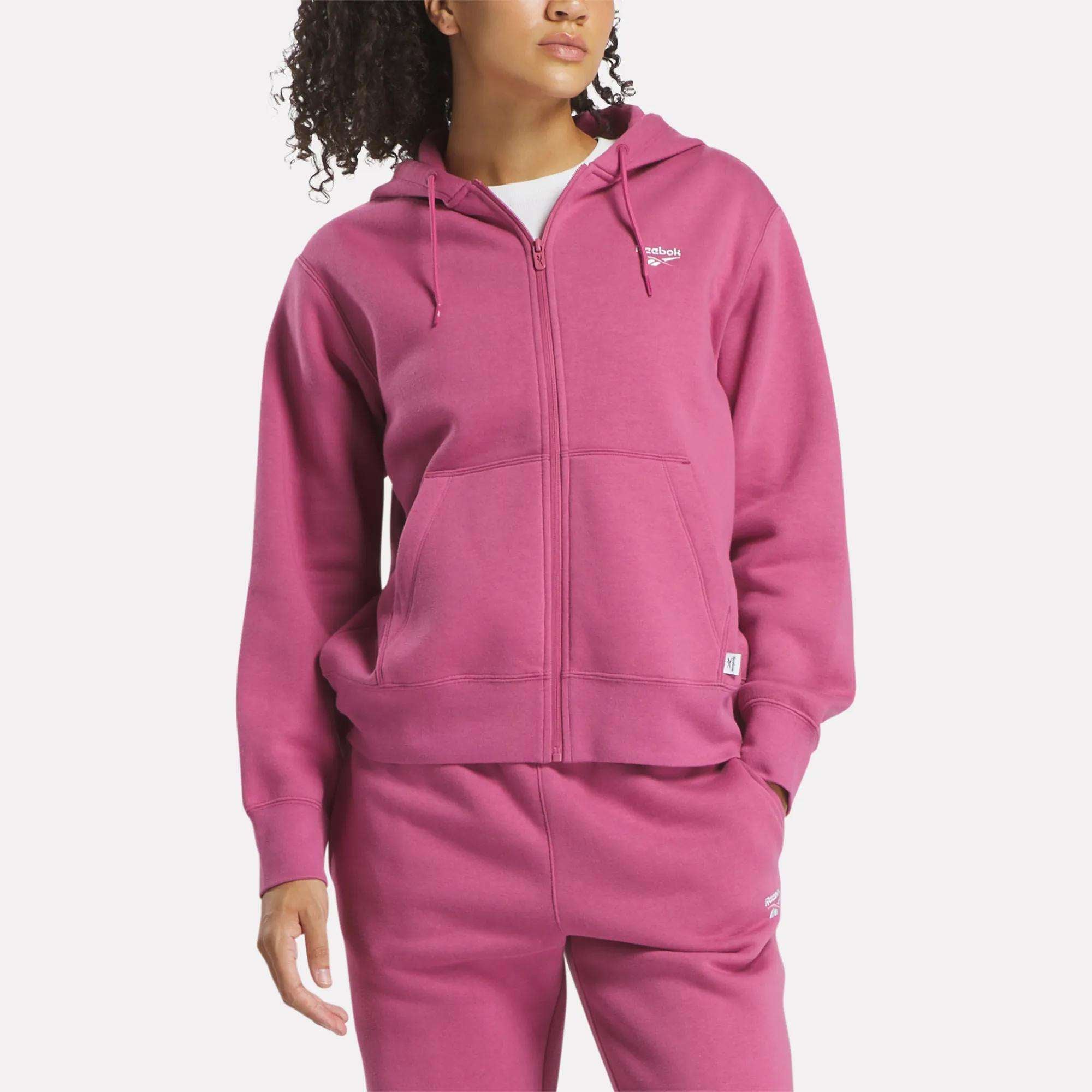 A woman wears the Reebok Women's Small Vector Full Zip Sweatshirt in magenta, paired with matching sweatpants. Both pieces display a white logo on the left chest and thigh as she stands against a plain white background.