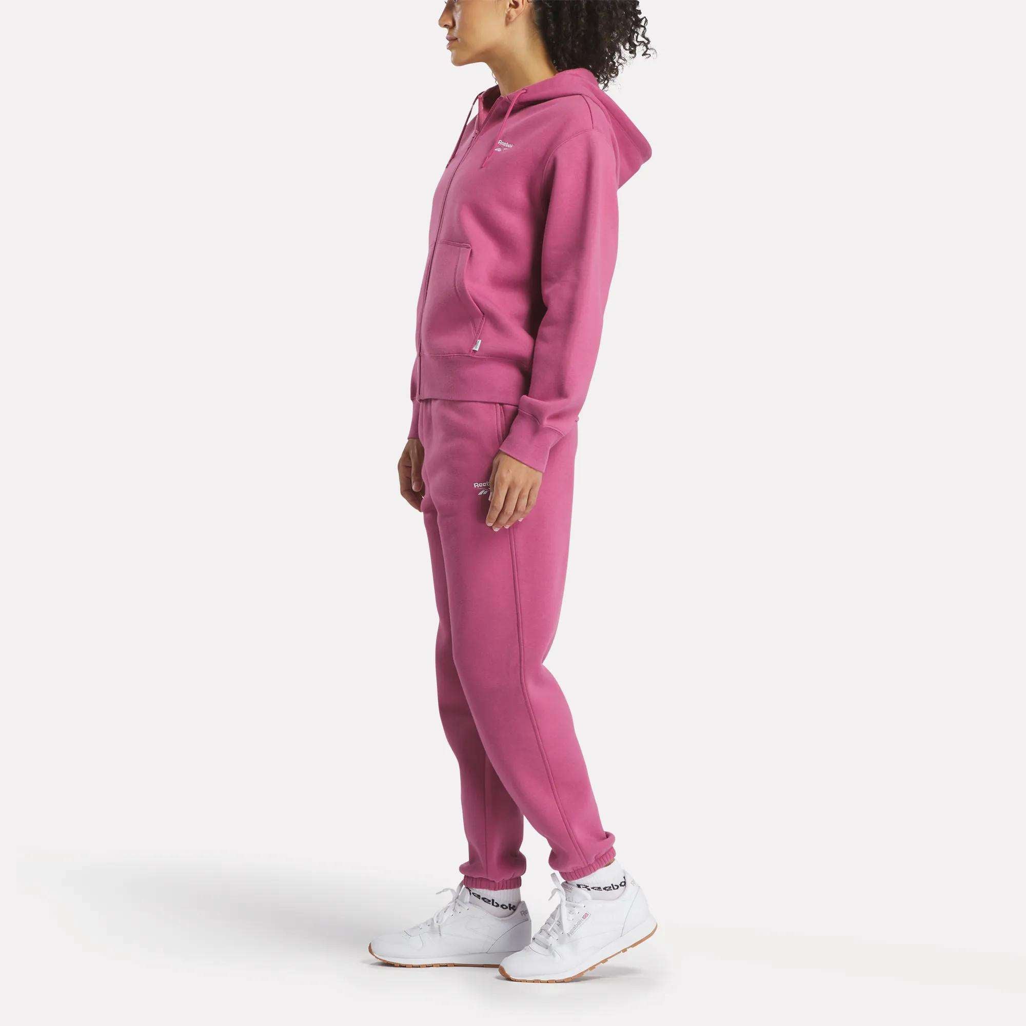 A woman stands in profile wearing a pink Reebok Women's Small Vector Full Zip Sweatshirt with matching sweatpants and white athletic shoes, set against a plain white background.