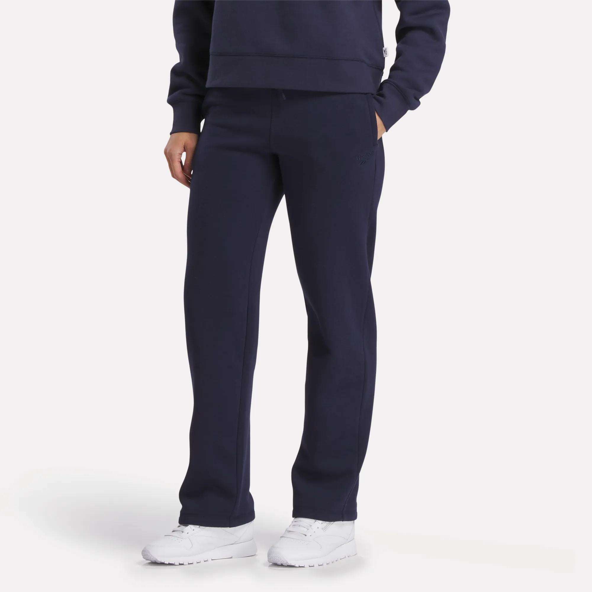 Only the lower half of the body is visible, showing a person in navy blue Reebok Women's Cozy Classic Sweatpants and a matching sweatshirt, both crafted from a soft cotton blend, paired with white sneakers against a plain light background.