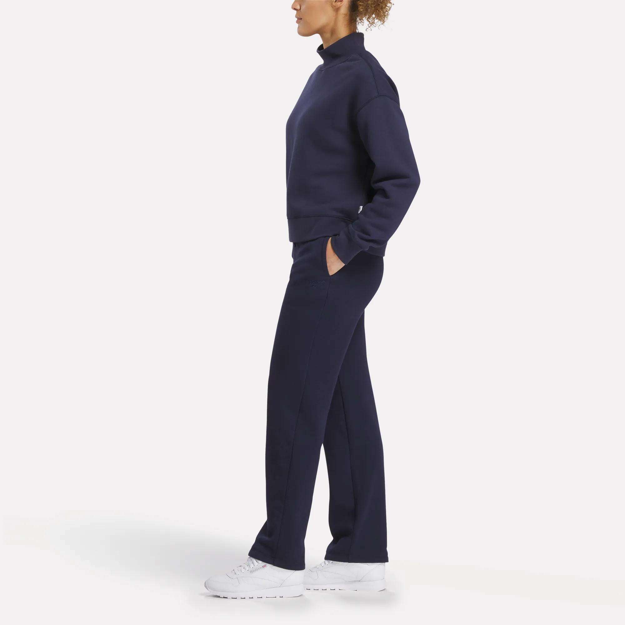 A person stands in profile wearing Reebok Women's Cozy Classic Sweatpants in navy blue, made from a soft cotton blend, with hands in pockets and paired with white sneakers, set against a plain white background.