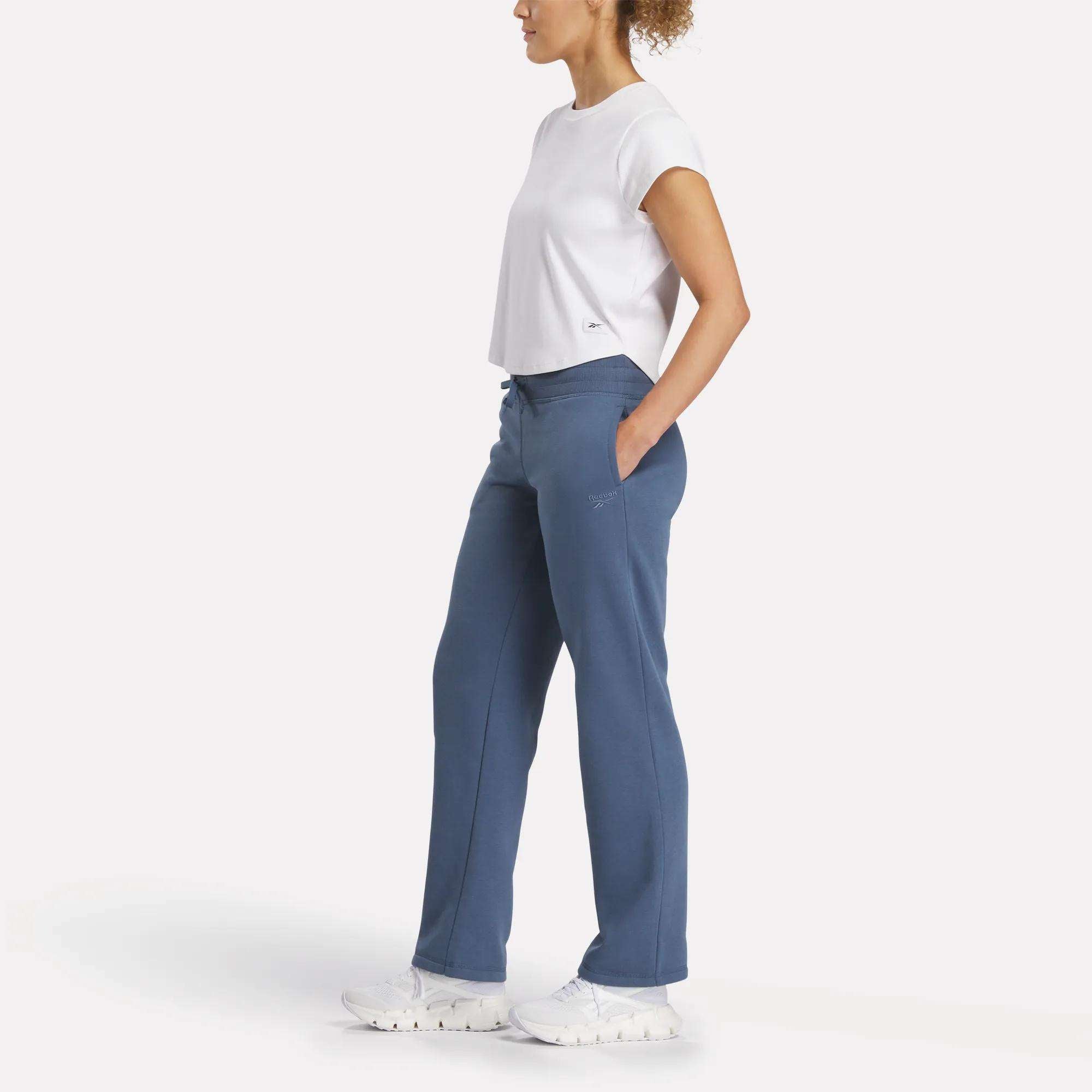 A person stands sideways, wearing a white short-sleeve shirt, white sneakers, and blue Reebok Women's Cozy Classic Sweatpants against a plain white background.