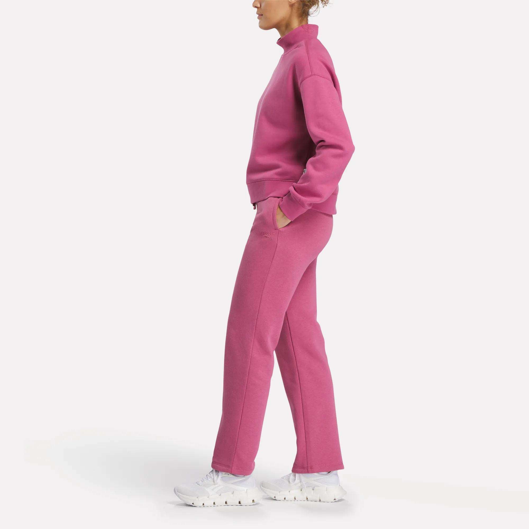 A woman stands in profile wearing Reebok's Women's Cozy Classic Sweatpants and a matching pink cotton-blend sweatshirt with white sneakers. Her hands are in her pockets, facing left against a plain white background.
