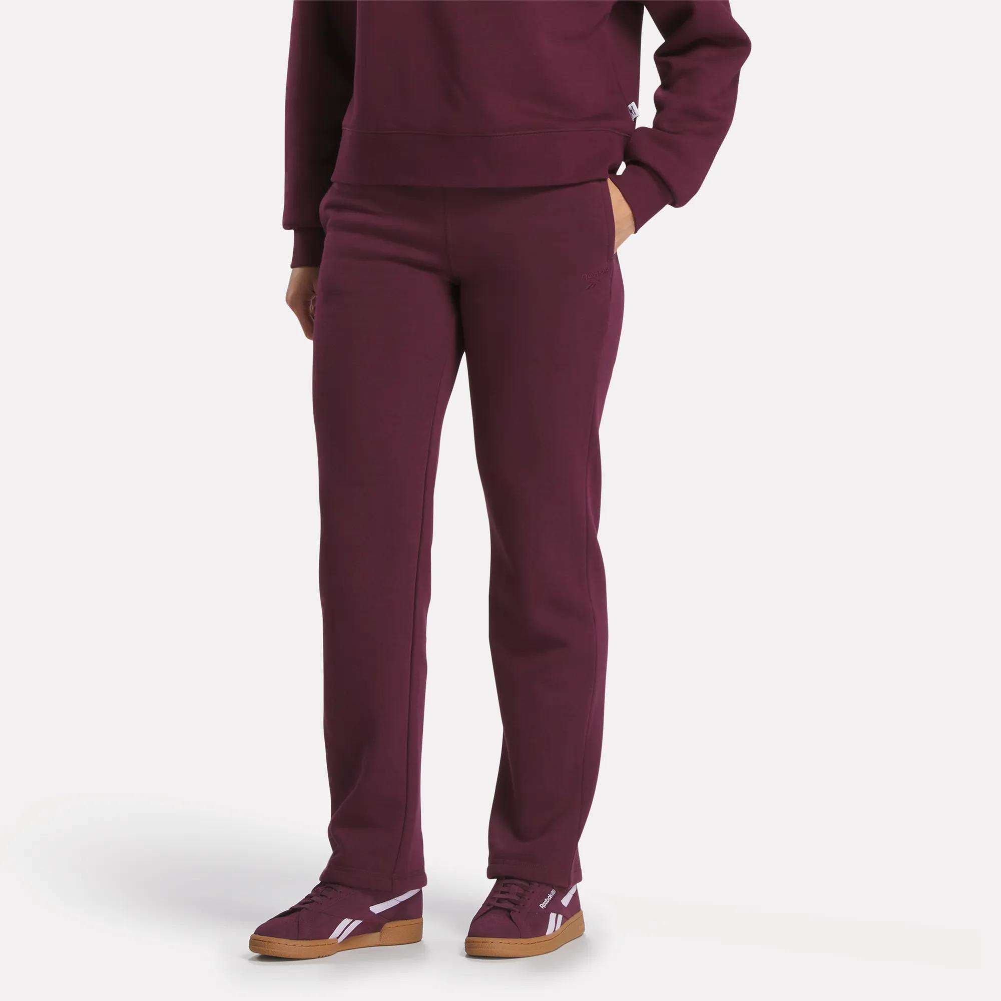 A person wearing Reebok Women's Cozy Classic Sweatpants in burgundy with a matching sweatshirt and burgundy sneakers with white accents stands with one hand in their pocket against a plain white background.